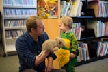 A man in a blue plaid shirt is kneeling on the floor of a library, engaging with a young child. The man holds a fluffy, gray puppet, and the child, wearing a green and yellow outfit, looks at him with a smile. Shelves filled with books and DVDs are in the background.