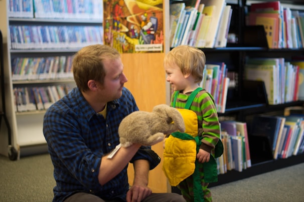A man in a blue plaid shirt is kneeling on the floor of a library, engaging with a young child. The man holds a fluffy, gray puppet, and the child, wearing a green and yellow outfit, looks at him with a smile. Shelves filled with books and DVDs are in the background.