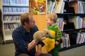 A man in a blue plaid shirt is kneeling on the floor of a library, engaging with a young child. The man holds a fluffy, gray puppet, and the child, wearing a green and yellow outfit, looks at him with a smile. Shelves filled with books and DVDs are in the background.