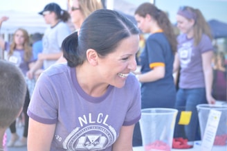 A woman with dark hair tied back is smiling and engaging with someone off-camera. She is wearing a purple shirt with an emblem and is outdoors, possibly at an event. Several other people are in the background, appearing to be participating in various activities. There are baskets and a table with some items on it.