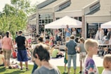 A lively outdoor event with people of all ages gathered on a grassy area in front of a building. A person is speaking into a microphone under a white canopy, while others stand nearby. Children and adults are scattered across the lawn, some seated and others standing, enjoying the sunny day. Several bubbles can be seen floating through the air.