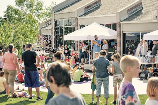 A lively outdoor event with people of all ages gathered on a grassy area in front of a building. A person is speaking into a microphone under a white canopy, while others stand nearby. Children and adults are scattered across the lawn, some seated and others standing, enjoying the sunny day. Several bubbles can be seen floating through the air.