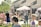 A lively outdoor event with people of all ages gathered on a grassy area in front of a building. A person is speaking into a microphone under a white canopy, while others stand nearby. Children and adults are scattered across the lawn, some seated and others standing, enjoying the sunny day. Several bubbles can be seen floating through the air.