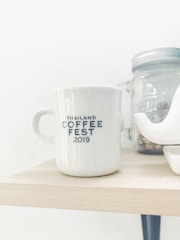 A white coffee mug featuring the text 'Thailand Coffee Fest 2019' is placed on a light wooden shelf. Next to it is a glass mason jar with a silver lid and a white ceramic object.