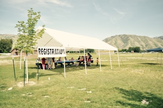 A registration tent set up in a grassy field with a clear sky and mountains in the background. A small group of people, including adults and children, are gathered under the white tent, which has a 'REGISTRATION' sign attached to it.