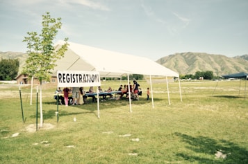 A registration tent set up in a grassy field with a clear sky and mountains in the background. A small group of people, including adults and children, are gathered under the white tent, which has a 'REGISTRATION' sign attached to it.