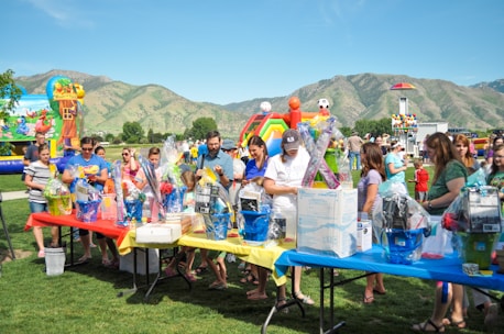 A group of people gathered around tables filled with gift baskets at an outdoor event. The tables are covered with vibrant red, yellow, and blue tablecloths. In the background, there are inflatable attractions, including a slide shaped like a colorful rainbow, soccer balls, and a beach-themed bounce house. The setting is a grassy park surrounded by green hills under a clear blue sky.