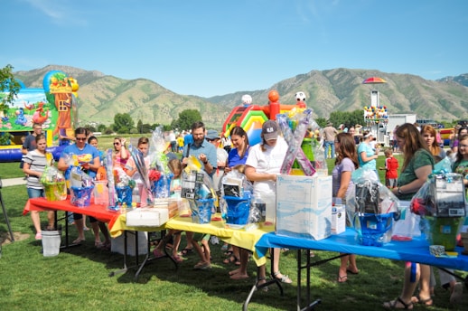A group of people gathered around tables filled with gift baskets at an outdoor event. The tables are covered with vibrant red, yellow, and blue tablecloths. In the background, there are inflatable attractions, including a slide shaped like a colorful rainbow, soccer balls, and a beach-themed bounce house. The setting is a grassy park surrounded by green hills under a clear blue sky.