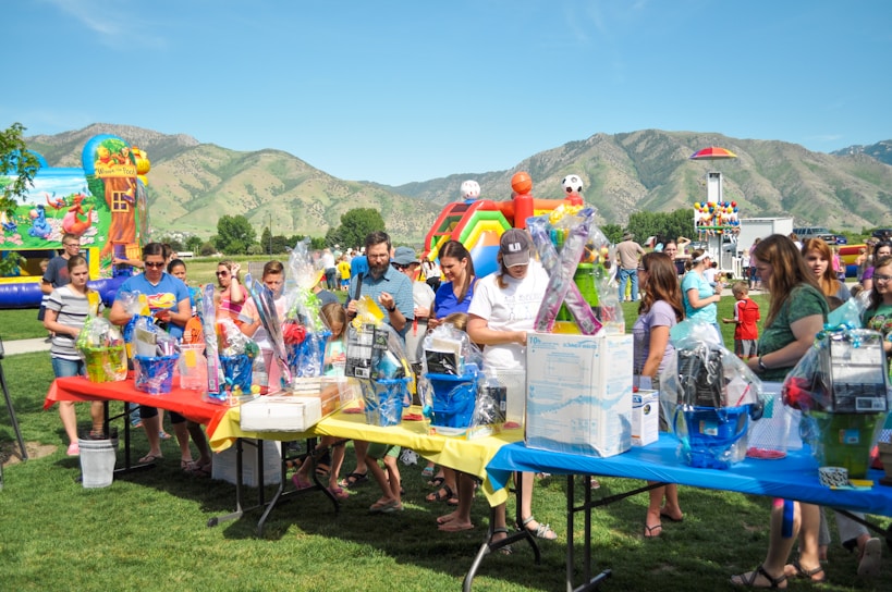 A group of people gathered around tables filled with gift baskets at an outdoor event. The tables are covered with vibrant red, yellow, and blue tablecloths. In the background, there are inflatable attractions, including a slide shaped like a colorful rainbow, soccer balls, and a beach-themed bounce house. The setting is a grassy park surrounded by green hills under a clear blue sky.
