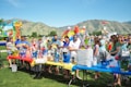 A group of people gathered around tables filled with gift baskets at an outdoor event. The tables are covered with vibrant red, yellow, and blue tablecloths. In the background, there are inflatable attractions, including a slide shaped like a colorful rainbow, soccer balls, and a beach-themed bounce house. The setting is a grassy park surrounded by green hills under a clear blue sky.