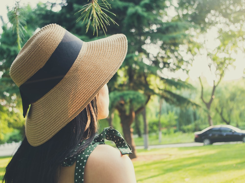 woman wearing hat standing near green field viewing black sedan parking surrounded with tall and green trees