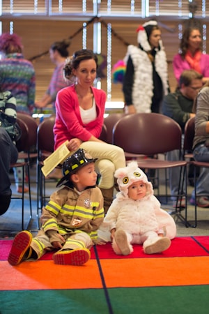 A lively gathering with children dressed in costumes, including a child in a firefighter outfit and a baby in a fluffy owl costume. They are seated on a colorful carpet, surrounded by adults who are either seated or standing in the background.