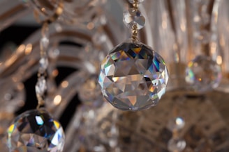 A close-up view of a sparkling crystal ornament hanging from a chandelier. The crystal displays a range of refracted colors, creating a rainbow effect across its facets. The background is blurred, putting emphasis on the clarity and shine of the crystal piece.