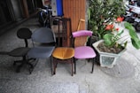 Four different styles of chairs are lined up side by side on an outdoor sidewalk. The chairs are in varying conditions and colors: one is black and wheeled, another is blue, an orange cushioned chair with a wooden backrest, and a purple chair with a curved seat. Next to the chairs, there is a large planter containing lush green plants and bright red flowers. Behind the chairs, there is a brick wall and several parked bicycles.