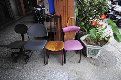 Four different styles of chairs are lined up side by side on an outdoor sidewalk. The chairs are in varying conditions and colors: one is black and wheeled, another is blue, an orange cushioned chair with a wooden backrest, and a purple chair with a curved seat. Next to the chairs, there is a large planter containing lush green plants and bright red flowers. Behind the chairs, there is a brick wall and several parked bicycles.