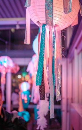 A vibrant indoor setting features hanging paper lanterns decorated with pink and teal ribbons. The lanterns emit a soft, glowing light, casting hues of pink, blue, and purple in the environment. The background is slightly blurred, suggesting a festive or celebratory atmosphere with people possibly present.