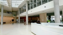 A modern, spacious lobby area with sleek architectural design. Large windows and glass walls allow natural light to illuminate the interior. There are concrete support pillars, a white reception desk, and a small potted plant visible. The flooring appears polished, contributing to a clean and professional atmosphere. Ceiling lights and structural patterns add to the contemporary feel.