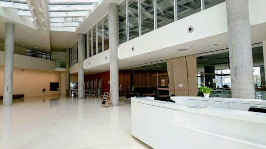 A modern, spacious lobby area with sleek architectural design. Large windows and glass walls allow natural light to illuminate the interior. There are concrete support pillars, a white reception desk, and a small potted plant visible. The flooring appears polished, contributing to a clean and professional atmosphere. Ceiling lights and structural patterns add to the contemporary feel.