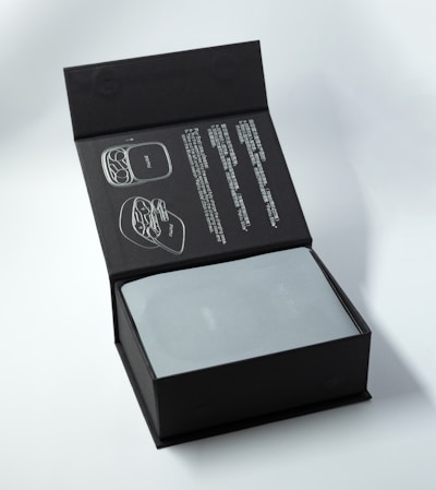 A black box with an instruction manual on the inside lid, featuring diagrams and text. The main compartment contains a metallic-looking object partially covered by a translucent material.