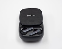 A pair of wireless earbuds sit in their charging case, which is open. The earbuds are sleek and modern in design and are resting inside the case. The case has a textured surface with a logo on top.
