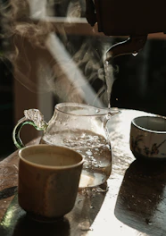 Hand pouring hot water from a kettle into a clear glass mug with herbal tea sachet inside.