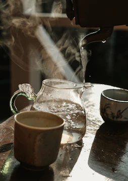Hand pouring hot water from a kettle into a clear glass mug with herbal tea sachet inside.