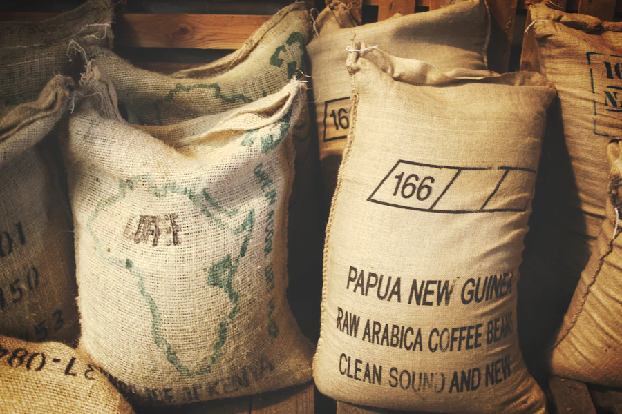 Jute sacks stacked against a wooden wall, stencilled with a lot number and the words raw arabica coffee beans.