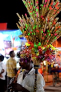 A person plays a flute while selling a colorful assortment of bamboo flutes and plastic toys, attached to a stick and carried on their shoulder. The background features blurred people and brightly illuminated stalls, suggesting a busy market or fair setting.