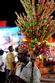 A person plays a flute while selling a colorful assortment of bamboo flutes and plastic toys, attached to a stick and carried on their shoulder. The background features blurred people and brightly illuminated stalls, suggesting a busy market or fair setting.