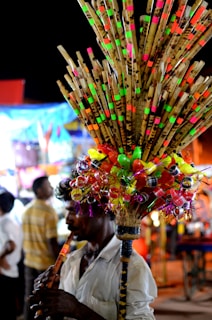 A person plays a flute while selling a colorful assortment of bamboo flutes and plastic toys, attached to a stick and carried on their shoulder. The background features blurred people and brightly illuminated stalls, suggesting a busy market or fair setting.