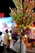 A person plays a flute while selling a colorful assortment of bamboo flutes and plastic toys, attached to a stick and carried on their shoulder. The background features blurred people and brightly illuminated stalls, suggesting a busy market or fair setting.