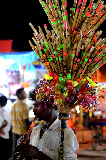 A person plays a flute while selling a colorful assortment of bamboo flutes and plastic toys, attached to a stick and carried on their shoulder. The background features blurred people and brightly illuminated stalls, suggesting a busy market or fair setting.