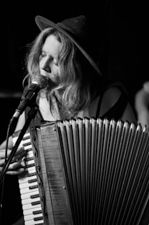 A person wearing a hat is playing an accordion and singing into a microphone. The image is in black and white, adding a classic and timeless feel. The focus is on the musician's expression and the details of the accordion.