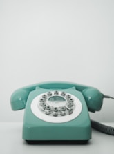 A vintage rotary dial telephone in a pastel teal color sits on a light surface. The phone has a classic circular dial with numbers and a coiled cord extending from the right side.