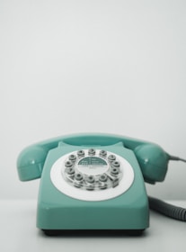 A vintage rotary dial telephone in a pastel teal color sits on a light surface. The phone has a classic circular dial with numbers and a coiled cord extending from the right side.