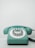 A vintage rotary dial telephone in a pastel teal color sits on a light surface. The phone has a classic circular dial with numbers and a coiled cord extending from the right side.