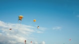 Various colorful kites of different shapes and sizes are flying against a clear blue sky with scattered clouds. One of the kites has a unique design resembling a face, and another appears to be shaped like a cartoon character.