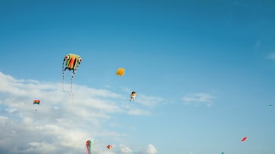 Various colorful kites of different shapes and sizes are flying against a clear blue sky with scattered clouds. One of the kites has a unique design resembling a face, and another appears to be shaped like a cartoon character.