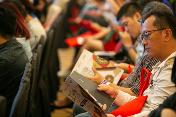 People seated in rows holding magazines or programs, appearing to be engaged in reading or browsing. They are facing forward, with some wearing glasses, and are holding red bags. The setting has a formal or organized event atmosphere.