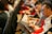 People seated in rows holding magazines or programs, appearing to be engaged in reading or browsing. They are facing forward, with some wearing glasses, and are holding red bags. The setting has a formal or organized event atmosphere.