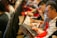 People seated in rows holding magazines or programs, appearing to be engaged in reading or browsing. They are facing forward, with some wearing glasses, and are holding red bags. The setting has a formal or organized event atmosphere.