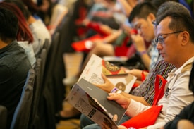 People seated in rows holding magazines or programs, appearing to be engaged in reading or browsing. They are facing forward, with some wearing glasses, and are holding red bags. The setting has a formal or organized event atmosphere.