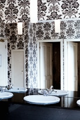 A bathroom features a monochrome floral wallpaper covering the walls, reflecting in the mirrors. Two white sinks with chrome faucets are mounted on a black countertop. Doors are visible in the background, adding to the sense of depth in the space.