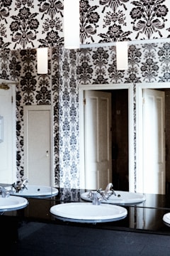 A bathroom features a monochrome floral wallpaper covering the walls, reflecting in the mirrors. Two white sinks with chrome faucets are mounted on a black countertop. Doors are visible in the background, adding to the sense of depth in the space.