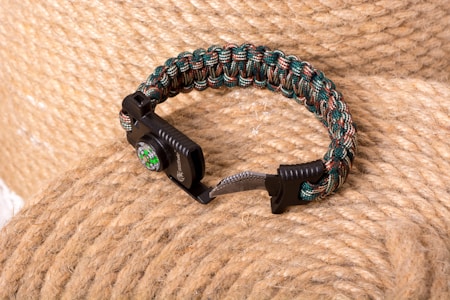 A braided survival bracelet with a built-in compass and a fire starter is placed on a coil of thick, coarse rope. The bracelet features a mix of green, brown, and black colors, and its rugged appearance suggests utility and outdoor usage.
