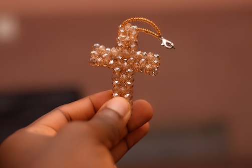 A hand holding a cross pendant made of translucent, beaded crystals. The pendant has a small clasp attached at the top, and the background is softly blurred, emphasizing the focus on the cross.