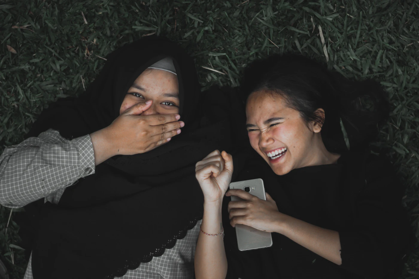 Dos amigas tumbadas en la hierba riéndose mirando un móvil