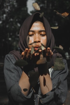 A person is gently blowing dry leaves from their hands, with their eyes closed and an expression of calm concentration. The background is blurred, creating a serene and focused scene.