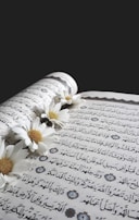 An open book with Arabic script is adorned with white daisies that are delicately placed along the center fold. The pages feature ornate decorative elements, enhancing the intricate text.