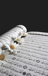 An open book with Arabic script is adorned with white daisies that are delicately placed along the center fold. The pages feature ornate decorative elements, enhancing the intricate text.